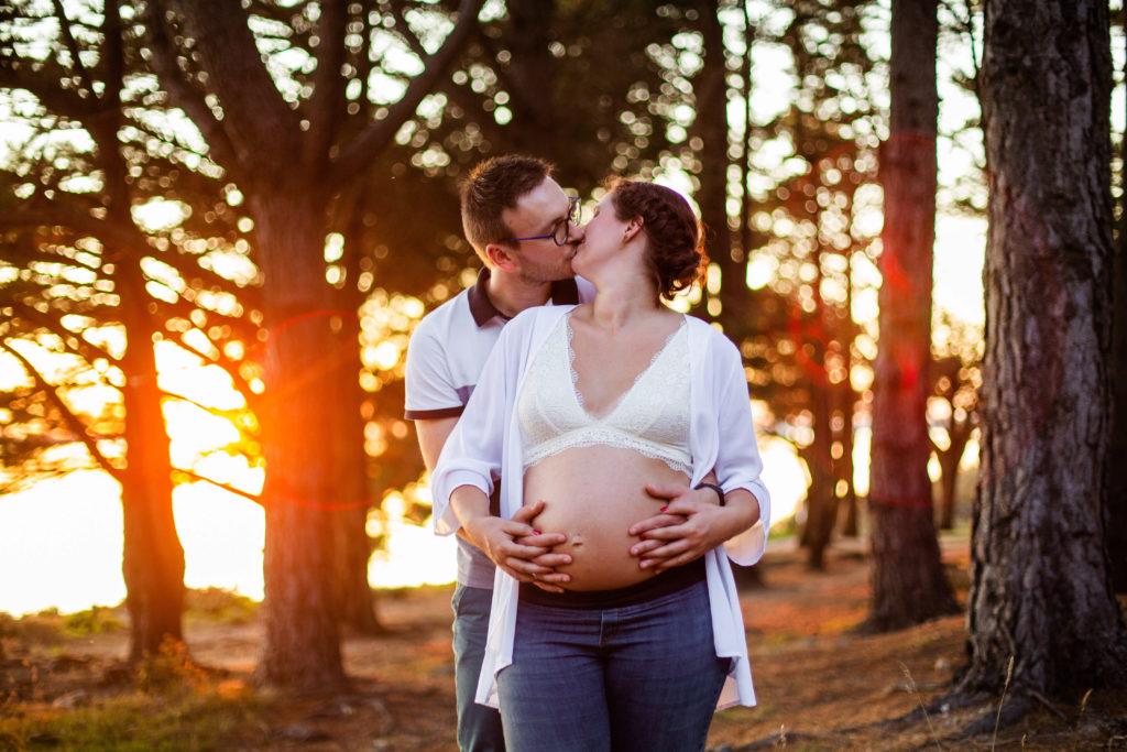 Couple s'embrassant dans une fôret pendant le coucher de soleil avec leur 4 mains sur le ventre de la femme